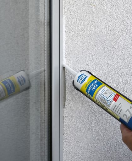 Eine Dichtmasse wird mit einer Kartuschenpistole auf eine Fensterrahmennaht aufgetragen. Die Wand ist hell und strukturiert, und die Dichtmasse wird gleichmäßig entlang der Kante aufgetragen. 