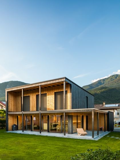 Modernes zweistöckiges Haus mit großen Fenstern und Holzfassade, in der Abenddämmerung beleuchtet. Das Haus ist von grünem Rasen und einem gepflegten Garten umgeben, im Hintergrund sind Berge und weitere Häuser zu sehen.