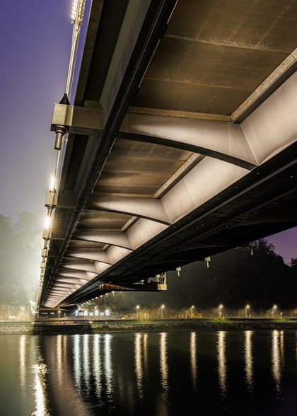 Eine nächtliche Luftaufnahme einer Brücke, die über einen Fluss führt. Die Brücke ist beleuchtet und reflektiert im Wasser.