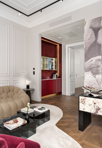 Wohnbereich der Opera Suite im „The Amauris“ Wien mit organisch geformtem beigen Sessel, gläsernen Couchtischen mit Bildbänden, Sideboard mit Steinfront, moderner Tischleuchte und tiefroter Nische mit beleuchteter Bar und Kaffeemaschine.