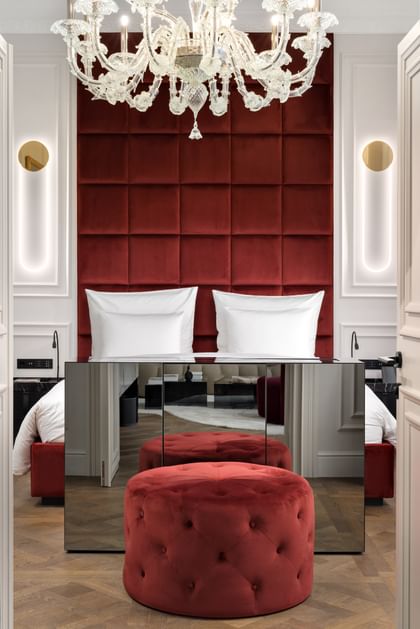 Elegantes Schlafzimmer der Opera Suite im „The Amauris“ Wien mit hohem bordeauxrotem Samtkopfteil hinter einem Doppelbett, großem Glaslüster, verspiegeltem Sideboard, rundem Samthocker und hellem Parkettboden.