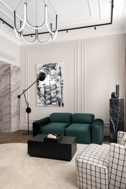 Moderner Sitzbereich im „The Amauris“ Wien mit grünem Samtsofa, hellem Teppich, abstrakter Wandkunst, skulpturaler Stehleuchte, schwarzem Couchtisch, kariertem Sessel und auffälligem, geschwungenen Deckenleuchten-Element.