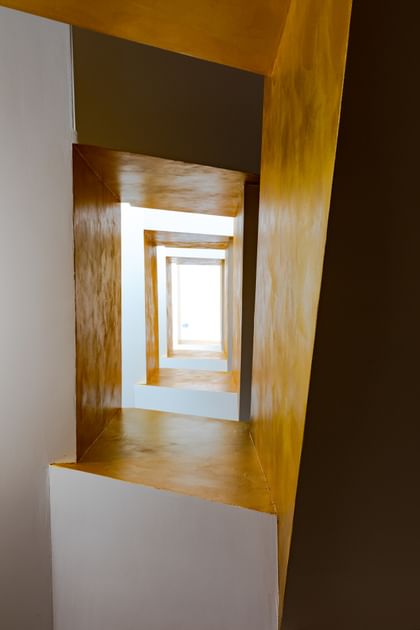 Eine perspektivische Ansicht eines langen, schmalen Raums mit Wänden in Weiß und Gelb. Die Wände sind in einem geometrischen Muster angeordnet, das einen Tiefeneffekt erzeugt. 