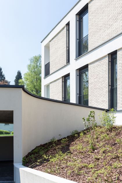Modernes Gebäude mit weißer und heller Ziegelfassade, großen Fenstern mit schwarzen Rahmen und einem überhängenden Bauteil. Im Vordergrund ein bepflanzter Hang, im Hintergrund Bäume und blauer Himmel.