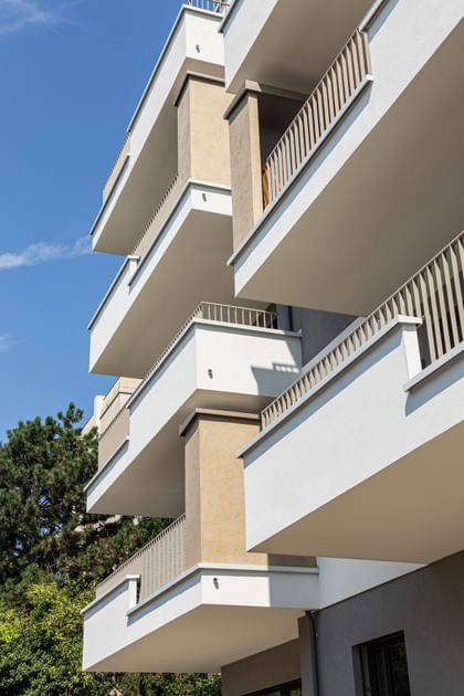 Wohnanlage in der Robert-Stolz-Straße in Linz. Ein modernes, mehrstöckiges Wohngebäude mit hellen Fassaden und zahlreichen Balkonen. Große Fenster sorgen für viel Tageslicht. Vor dem Gebäude stehen Autos auf einer ruhig wirkenden Straße, eingerahmt von gepflegten Grünflächen und Bäumen.