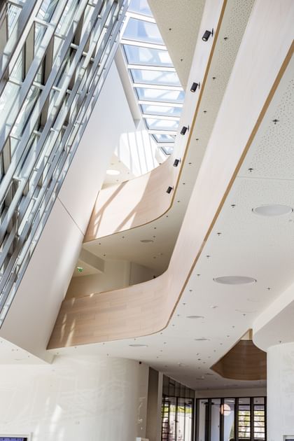 Großzügig gestalteter, lichtdurchfluteter Eingangsbereich der Anton Bruckner Privatuniversität mit imposanter Glasfassade, fließenden Holzbalkonen und moderner weißer Innenarchitektur; offene Atmosphäre und klare Linien prägen das Foyer.