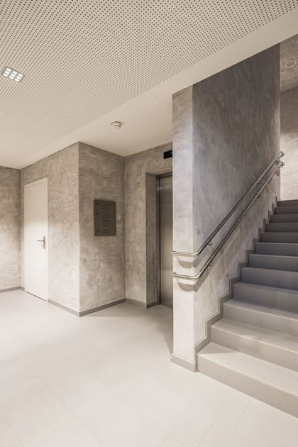 Flur mit Wänden in Betonoptik und einer Treppe, die in die weiteren Geschosse führt sowie ein Fahrstuhleingang