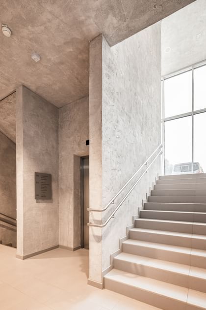 Flur mit Wänden in Betonoptik und einer Treppe, die in die weiteren Geschosse führt sowie ein Fahrstuhleingang