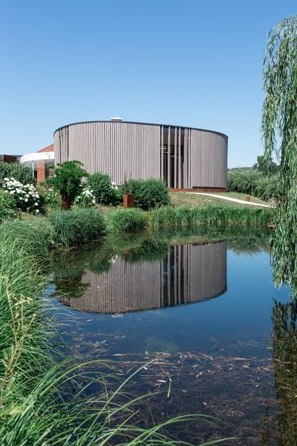 Rundes Gebäude mit einer Holzfassade spiegelt sich in einem ruhigen Teich, umgeben von üppigem Grün und blühenden Pflanzen