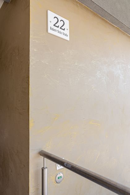 Wohnanlage in der Robert-Stolz-Straße in Linz. Ein modernes, mehrstöckiges Wohngebäude mit hellen Fassaden und zahlreichen Balkonen. Große Fenster sorgen für viel Tageslicht. Vor dem Gebäude stehen Autos auf einer ruhig wirkenden Straße, eingerahmt von gepflegten Grünflächen und Bäumen.