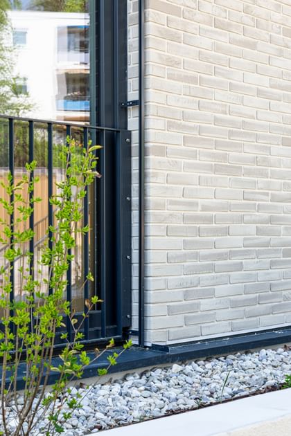  Nahaufnahme einer Gebäudefassade mit hellen Ziegeln, schwarzen Fensterbänken und Metallgittern an den Fenstern. Unten und rechts ist weißer Putz zu sehen, entlang der Wand verläuft ein Streifen mit weißen Kieselsteinen.