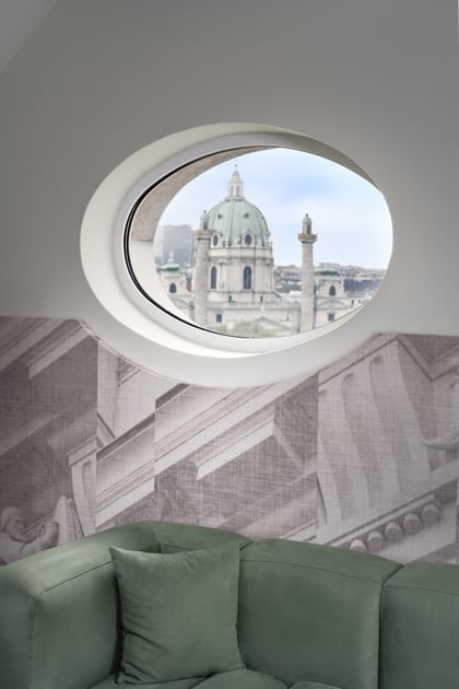 Loft-Suite im „The Amauris“ Wien mit rundem Bullaugenfenster und Blick auf die barocke Kuppel und Säulen der Karlskirche, davor eine grüne Samt-Eckcouch vor einer Wand mit großflächiger Architekturzeichnung.