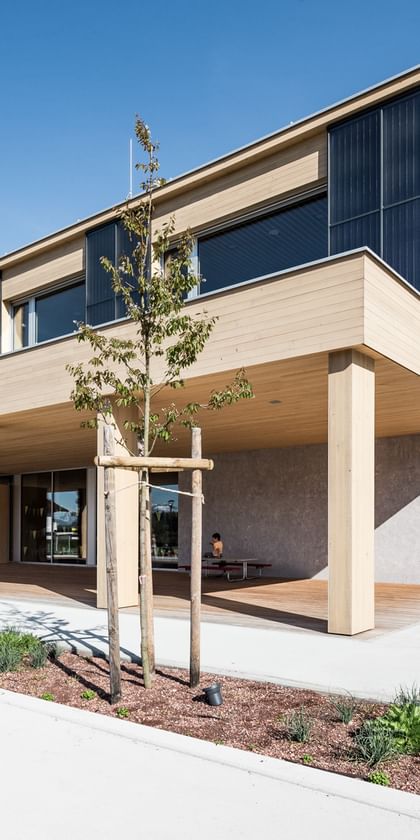 Holzfassade der Volkshochschule mit großen Fenstern. Vor dem Gebäude befinden sich einige Bäume und Sträucher.