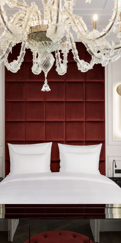 Schlafzimmer der Opera Suite im „The Amauris“ Wien mit großem Doppelbett vor hohem, weinrotem Kopfteil, vertikalen Lichtleisten in weißen Wandpanelen, opulentem Glaslüster und Einblick in ein marmorverkleidetes Badezimmer.