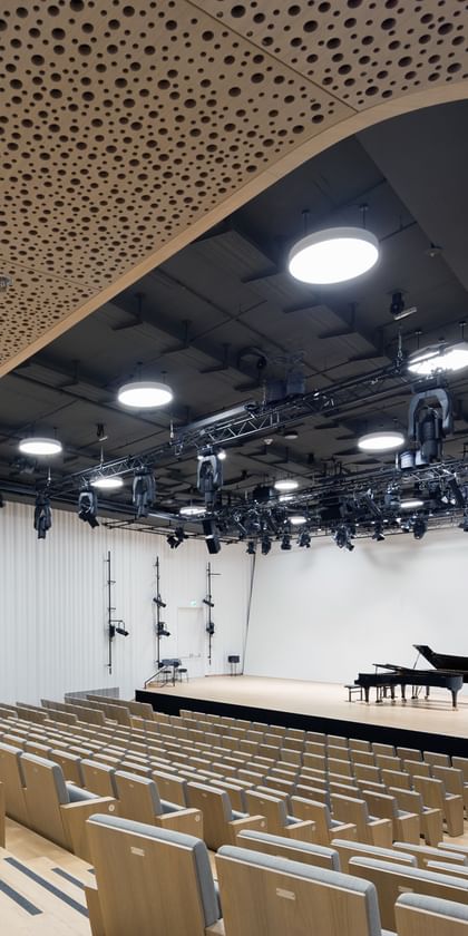 Blick von hinten in einen modernen Konzertsaal der Anton Bruckner Privatuniversität mit ansteigenden Sitzreihen, offen gestalteter Bühne und Flügel; hochwertige Akustikdecken, dezente Beleuchtung und klar strukturierte Architektur sorgen für ein helles, freundliches Ambiente.