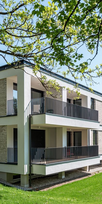 Modernes zweistöckiges Gebäude mit großen Fenstern und Balkonen, gelegen in einer grünen Umgebung mit Bäumen im Hintergrund bei sonnigem Wetter und klarem Himmel.