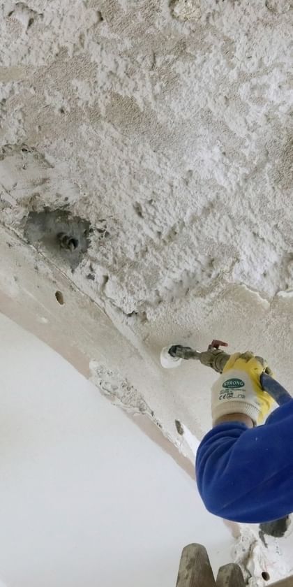 Ein Handwerker in blauer Jacke und eine schwarzer Mütze arbeitet mit einem Werkzeug an der Decke, die mit Putz und Bohrlöchern versehen ist.