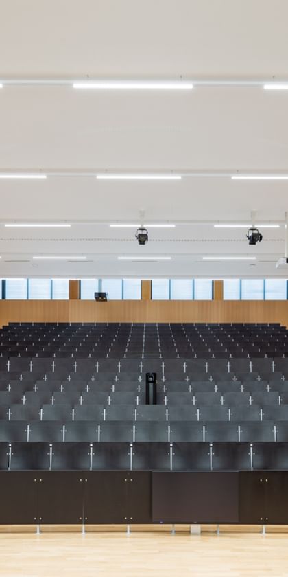 Leerer Hörsaal mit gestuften Sitzreihen, einem Rednerpult mit elektronischer Ausstattung und dekorativen Akustikpaneelen in verschiedenen Formen und Farben an den Holzverkleidungen.