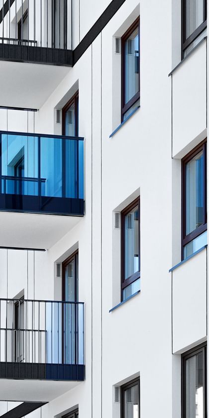 Ein mehrstöckiges Wohngebäude mir einer weißen Fassade. Es gibt zahlreiche Balkone mit schwarzen Geländern. Einige Balkone sind mit blauen Glaswänden ausgestattet.