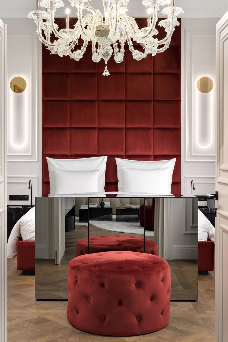 Elegantes Schlafzimmer der Opera Suite im „The Amauris“ Wien mit hohem bordeauxrotem Samtkopfteil hinter einem Doppelbett, großem Glaslüster, verspiegeltem Sideboard, rundem Samthocker und hellem Parkettboden.