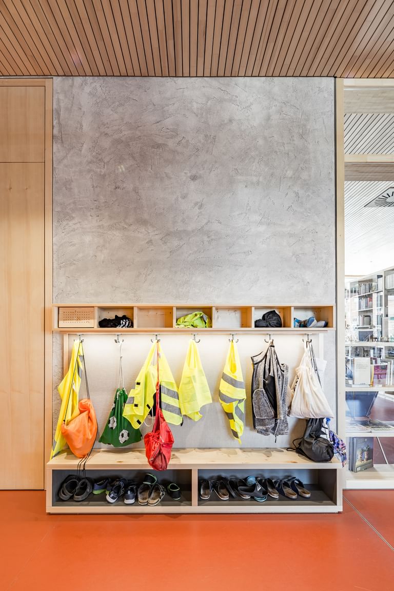Eingangsbereich einer Schule. An der Wand hängen mehrere leuchtend gelbe und grüne Warnwesten und Rücksäcke auf Kleiderhaken. Darunter befindet sich ein offenes Regal, in dem Schuhe verstaut sind.