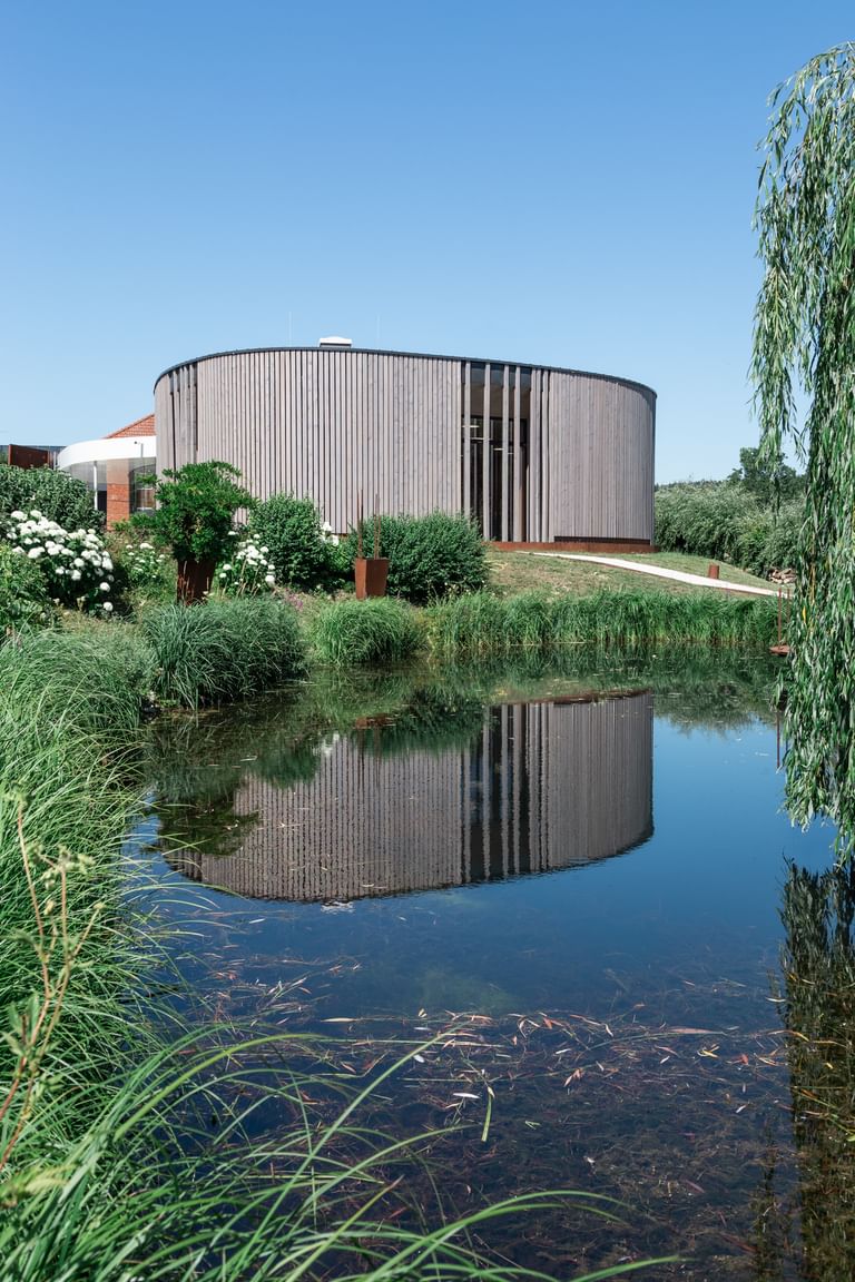 Rundes Gebäude mit einer Holzfassade spiegelt sich in einem ruhigen Teich, umgeben von üppigem Grün und blühenden Pflanzen