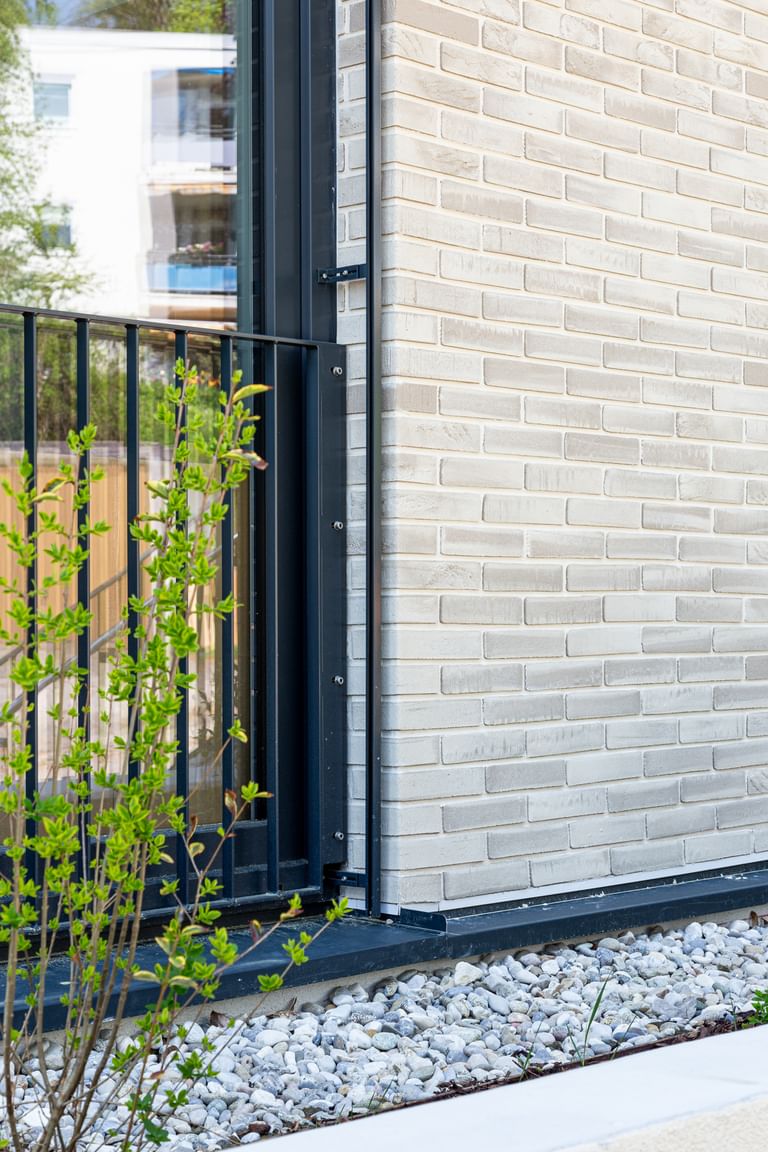 Nahaufnahme einer Gebäudefassade mit hellen Ziegeln, schwarzen Fensterbänken und Metallgittern an den Fenstern. Unten und rechts ist weißer Putz zu sehen, entlang der Wand verläuft ein Streifen mit weißen Kieselsteinen.