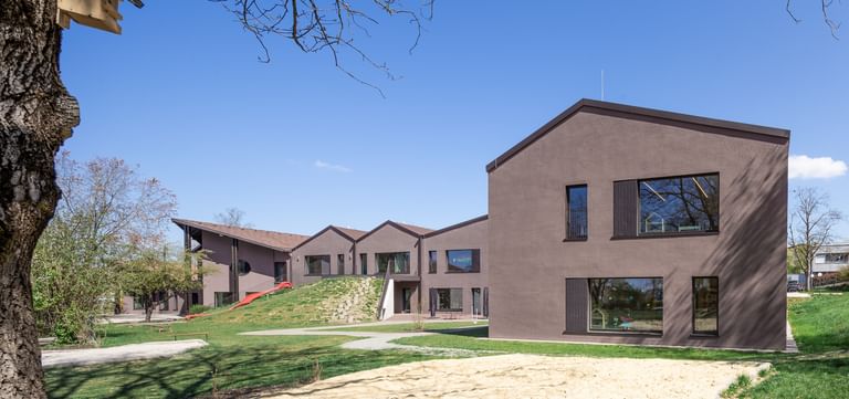 Zweigeschossiges Gebäude mit brauner Fassade und großen Fenstern, umgeben von einer grünen Wiese und Bäumen. Im Vordergrund ist ein Sandbereich sichtbar.