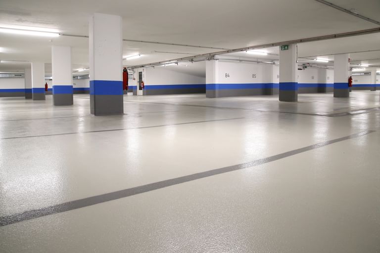 Ein leerer Parkhausbereich mit hellen Wänden und einem grauen Boden. Die Wände sind teilweise mit blauen Streifen dekoriert. Es gibt mehrere Säulen und einige Feuerlöscher an den Wänden.