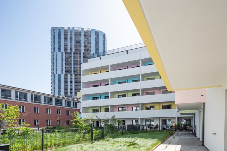 Mehrstöckiges Wohnhaus mit bunter Fassade, weißen Balkonen, Rasen und Bäumen, Hochhaus im Hintergrund