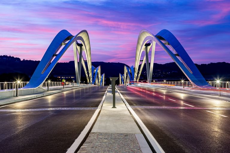 Brücke mit geschwungenen, weißen Bögen, die sich über eine Straße erstreckt. Der Himmel ist in sanften Lila- und Rosatönen gefärbt.