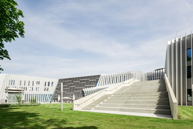 Modernes Gebäude der Anton Bruckner Privatuniversität mit markanter Fassadengestaltung aus vertikalen und horizontalen Lamellen. Eine breite Treppe führt zum Eingang, im Vordergrund befindet sich eine gepflegte Grünfläche.