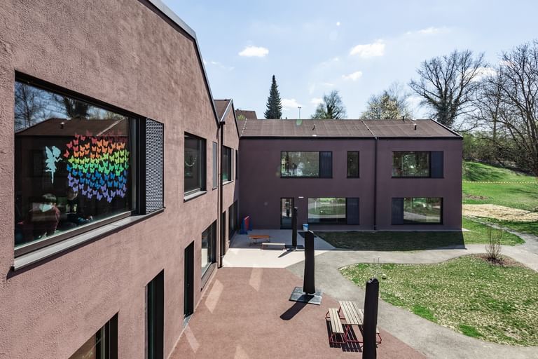 Zweigeschossiges Gebäude mit großen Fenstern und schwarzen Fensterläden. In den oberen Fenstern sind bunte Zeichnungen sichtbar.