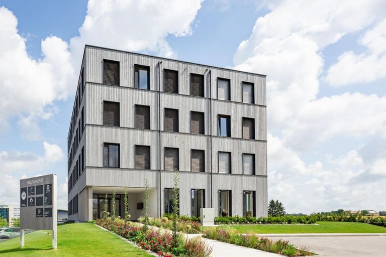 Ein mehrstöckiges Gebäude mit einer Fassade aus Holz und großen Fenstern. Es hat eine rechteckige Form und ist von einem gepflegten Garten mit Blumen und Sträuchern umgeben