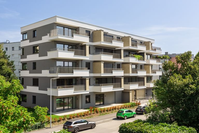 Wohnanlage in der Robert-Stolz-Straße in Linz. Ein modernes, mehrstöckiges Wohngebäude mit hellen Fassaden und zahlreichen Balkonen. Große Fenster sorgen für viel Tageslicht. Vor dem Gebäude stehen Autos auf einer ruhig wirkenden Straße, eingerahmt von gepflegten Grünflächen und Bäumen.