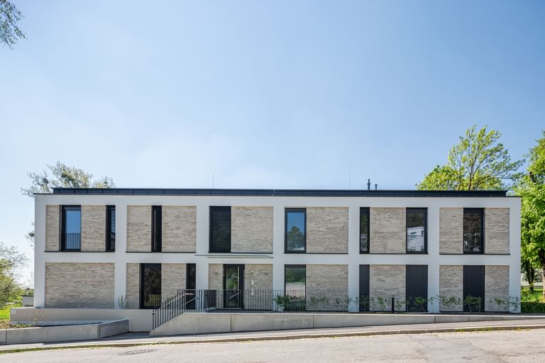 Modernes zweistöckiges Gebäude mit großen Fenstern und Balkonen, mit Bäumen im Hintergrund bei sonnigem Wetter und klarem Himmel.