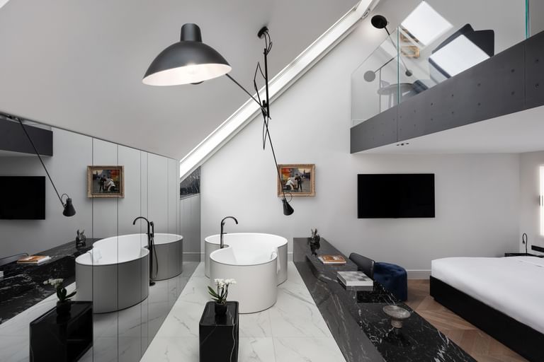 Loftartige Grand-Maisonette-Suite im „The Amauris“ Wien mit zwei freistehenden runden Badewannen auf hellem Marmorboden, langen schwarzen Marmorablagen mit Deko, großem Doppelbett und Galerieebene mit Glasbrüstung unter schräger Dachdecke.