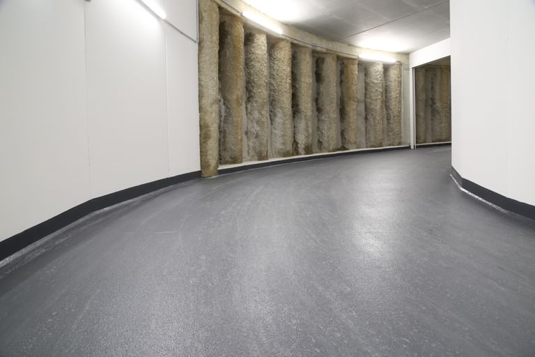 Tiefgarage mit einem gewundenen, grauen Boden und einer Wand, die aus vertikalen, strukturierten Elementen besteht. Die Wände sind weiß und die Beleuchtung ist gleichmäßig.