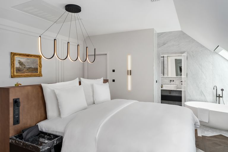 Modernes Schlafzimmer der Loft Suite im „The Amauris“ Wien mit großem Doppelbett, Holz-Kopfteil, organischer schwarzer Deckenleuchte, Wandgemälde und Blick in ein helles Badezimmer mit freistehender Badewanne und Marmorflächen.