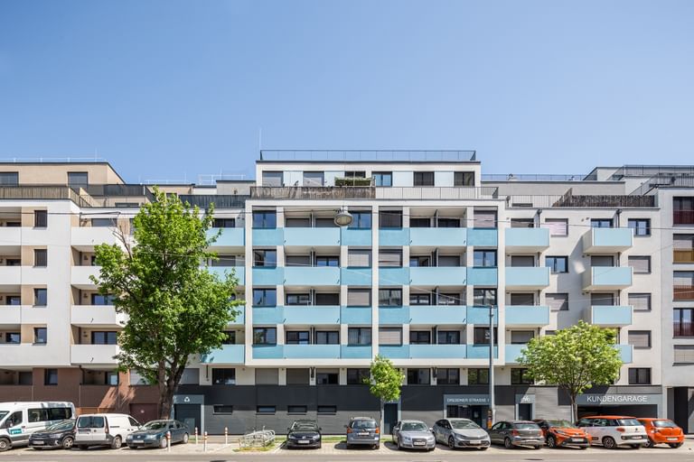 Straßenansicht der Fassade eines Mehrparteienhauses, blaue Balkone, davor parken Autos