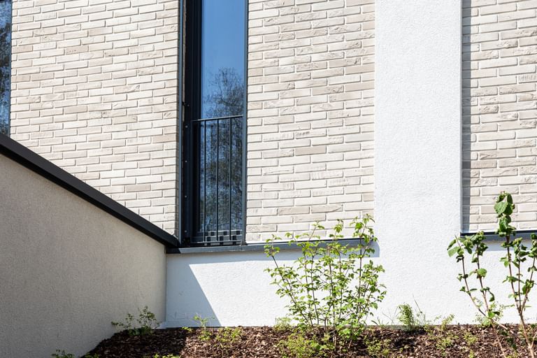  Nahaufnahme einer Gebäudefassade mit hellen Ziegeln, schwarzen Fensterbänken und Metallgittern an den Fenstern. Unten und rechts ist weißer Putz zu sehen, entlang der Wand verläuft ein Streifen mit weißen Kieselsteinen.