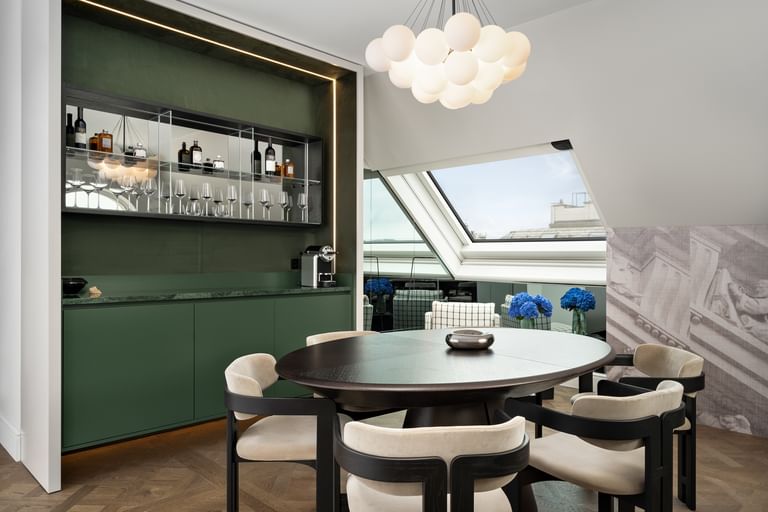 Ess- und Loungebereich der Loft Suite im „The Amauris“ Wien mit rundem Holztisch und vier gepolsterten Stühlen, eleganter grüner Einbau-Bar mit Spiegelregal, Flaschen und Gläsern sowie großen Dachfenstern mit Stadtblick und blauen Hortensien.