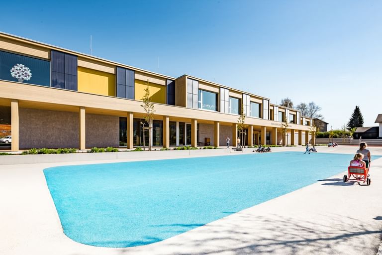  Außenansicht der Volkshochschule. Im Vordergrund sind zwei Kinder auf einem roten Spielzeugfahrzeug zu sehen, die vor einer blauen Spielfläche spielen, während andere Kinder mehr im Hintergrund spielen