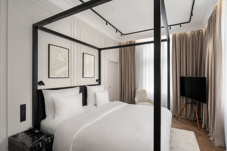 Helles Executive-Suite-Schlafzimmer im „The Amauris“ Wien mit modernem Himmelbett in schwarzem Rahmen, weißer Bettwäsche, abstrakten Wandbildern, bodentiefen Vorhängen, Lounge-Sessel und freistehendem TV auf hellem Parkett.