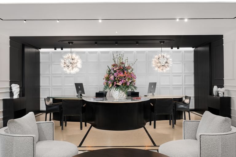 Helle Hotellobby des „The Amauris“ Wien mit halbkreisförmiger dunkler Rezeptionstheke, großem Blumenarrangement, drei Arbeitsplätzen vor kassettierten weißen Wänden, zwei kugelförmigen Glasleuchten und zwei gepolsterten Lounge-Sesseln im Vordergrund.