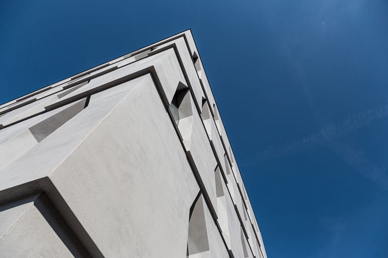 Die Ecke eines Gebäudes, die in die Höhe ragt. Die Fassade ist aus hellem Beton mit schmalen Fenstern, vor einem klaren blauen Himmel.