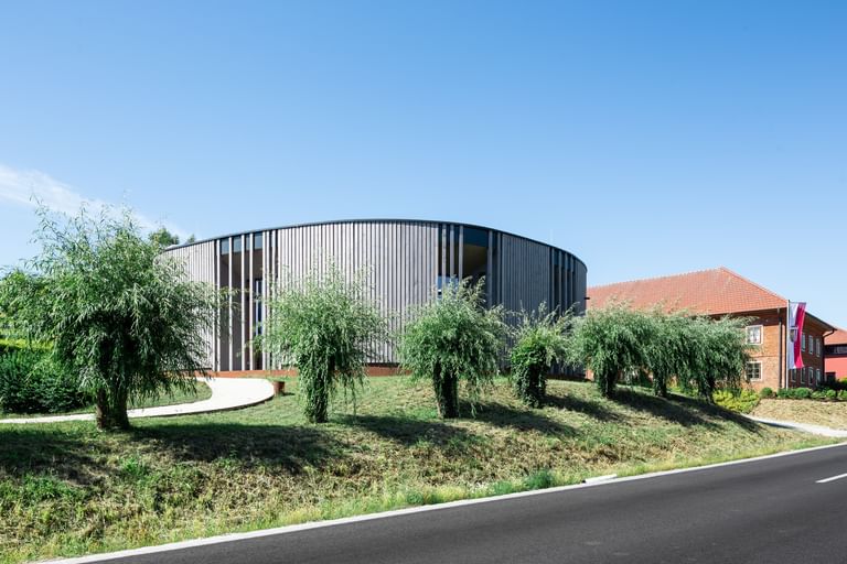 Rundes Gebäude mit einer Holzfassade, das von einer grünen Landschaft umgeben ist. Im Vordergrund sind mehrere Sträucher oder kleine Bäume zu sehen