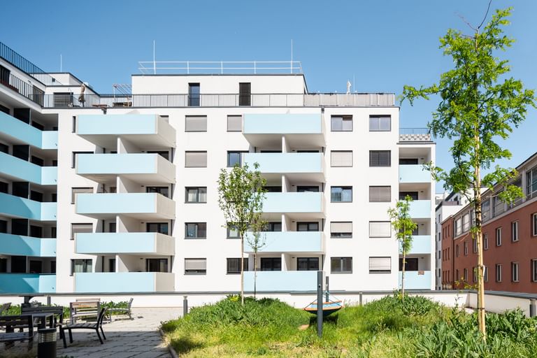 Mehrstöckiges Wohnhaus mit weißer Fassade, türkisen Balkonen, Rasen und Bäumen