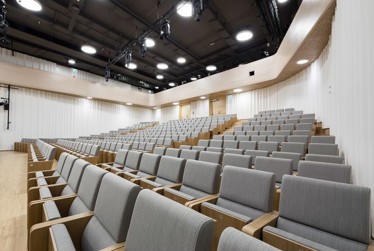 Moderner, großzügiger Hörsaal der Anton Bruckner Privatuniversität mit ansteigenden, gepolsterten Holzsitzreihen und hell gestalteter, wellenförmiger Wandverkleidung; ausgestattet mit moderner Deckenbeleuchtung und Veranstaltungstechnik – ideal für Vorträge, Konferenzen oder Präsentationen.
