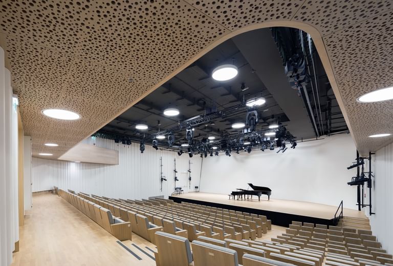 Blick von hinten in einen modernen Konzertsaal der Anton Bruckner Privatuniversität mit ansteigenden Sitzreihen, offen gestalteter Bühne und Flügel; hochwertige Akustikdecken, dezente Beleuchtung und klar strukturierte Architektur sorgen für ein helles, freundliches Ambiente.